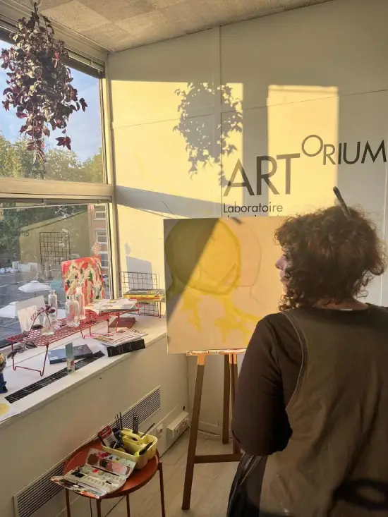 Animatrice peignant un tableau assise sur un tabouret, vue de dos, dans l’atelier de l'Artorium baigné de lumière
